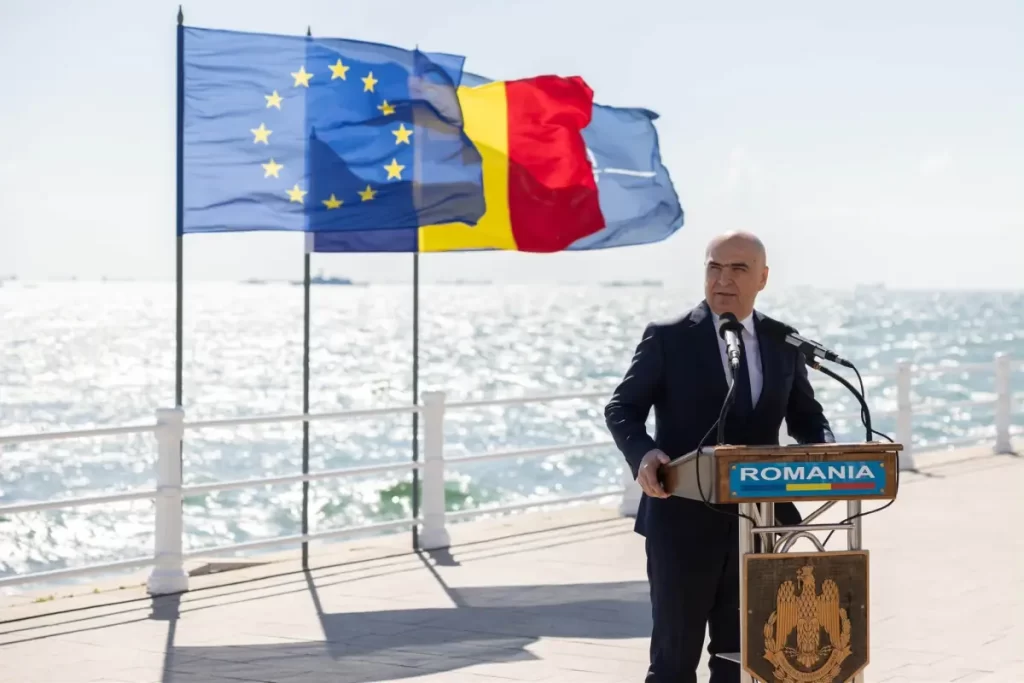 CAP BOLOJAN Romanian official giving a speech at a seaside podium with EU and Romania flags in the background.