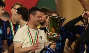 Man in a light shirt holding a large gold trophy while surrounded by teammates wearing blue jerseys and medals.