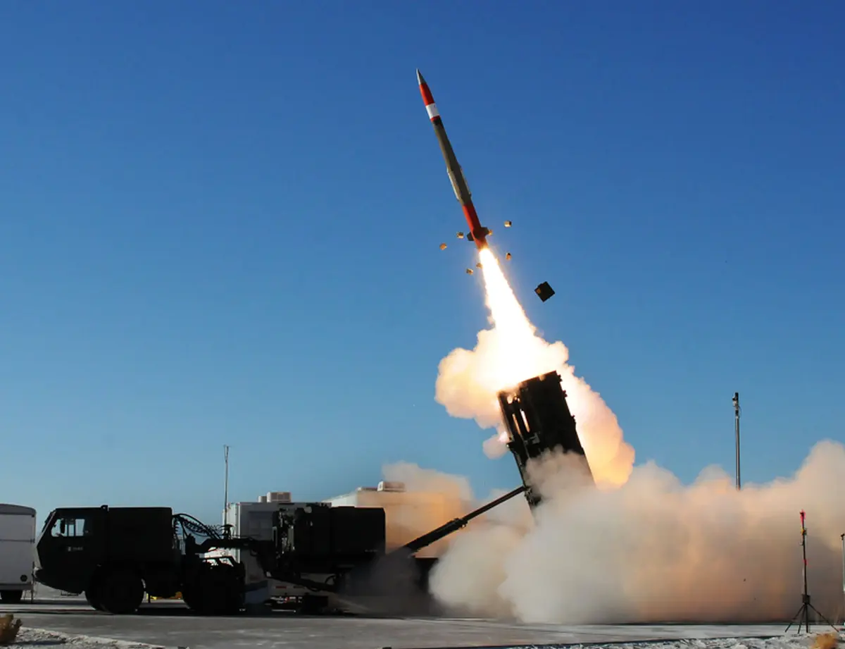 Rocket launching from ground-based launcher with bright flame and smoke plume against a blue sky, support vehicles visible nearby as the rocket rises.