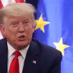 Close-up of a man in a dark suit and red tie speaking, with US and EU flags in the background.
