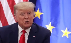 Close-up of a man in a dark suit and red tie speaking, with US and EU flags in the background.