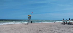 Lifeguard tower with a red flag on a sandy beach, blue ocean waves, and thatched umbrellas with people relaxing nearby.