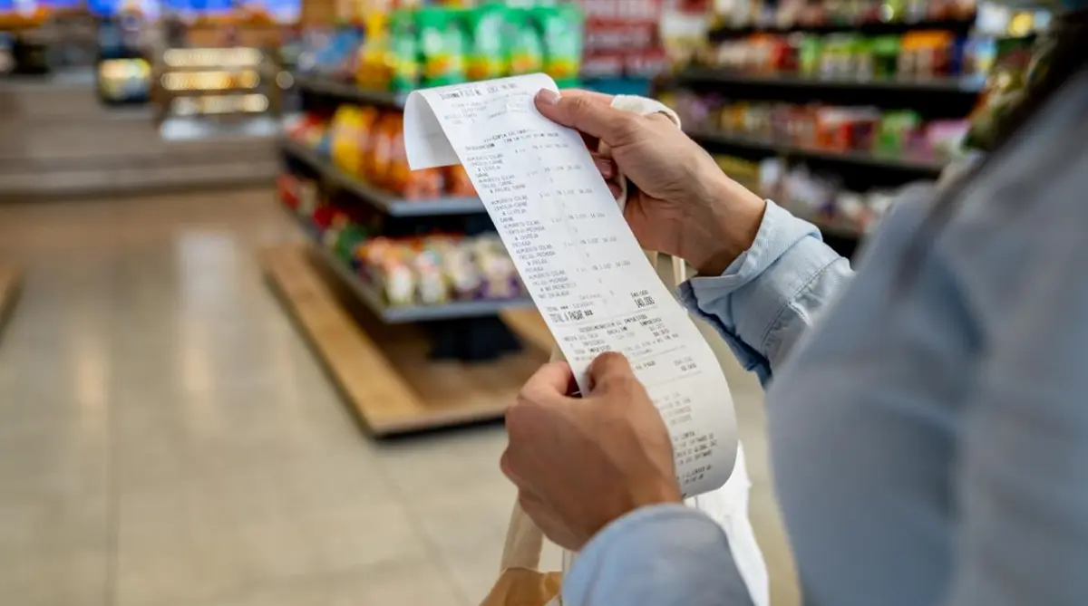 Person in a blue shirt holds a long grocery receipt in a supermarket, shelves blurred in the background.
