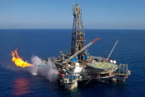 Offshore oil rig with a large flare stack burning gas, flames and water spray surrounding the platform in the ocean.